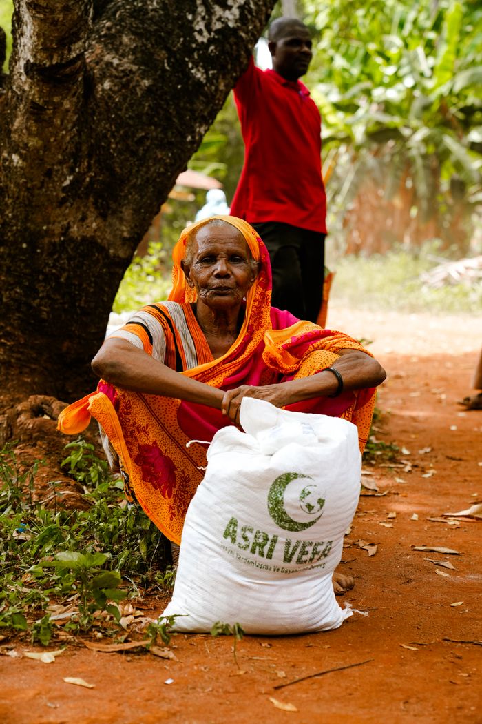 Elderly woman sits by tree with a relief bag labeled 'ASRI VEER'. Rural outdoor setting.