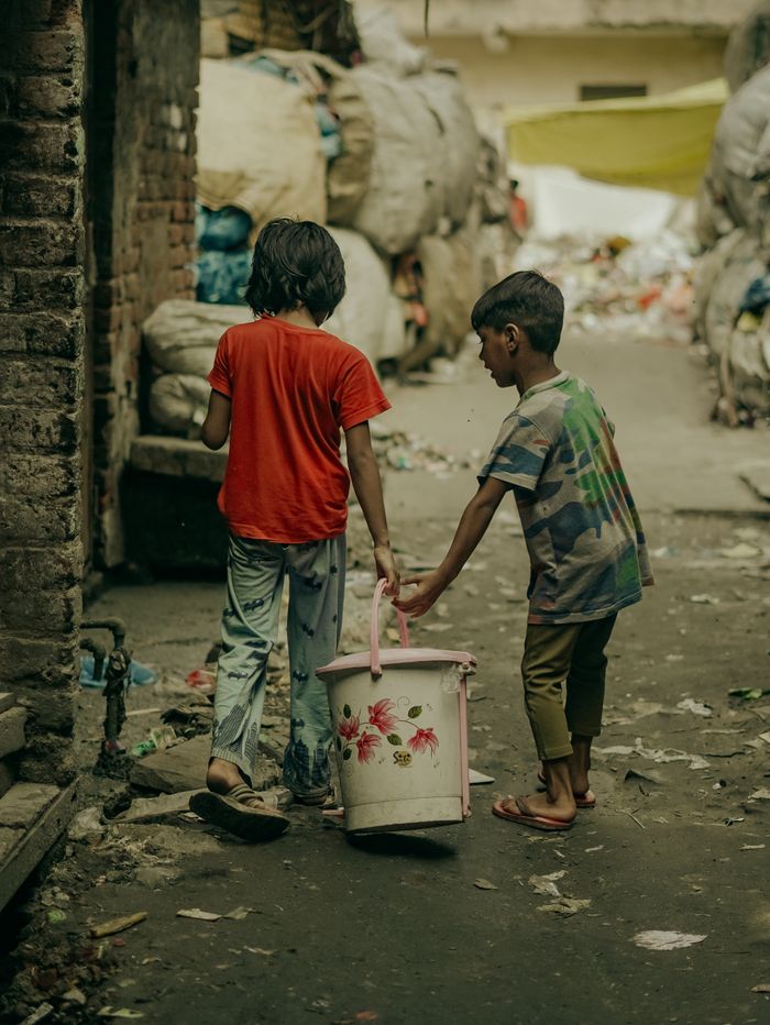 Two children in a New Delhi alley carrying a water bucket, highlighting urban life.