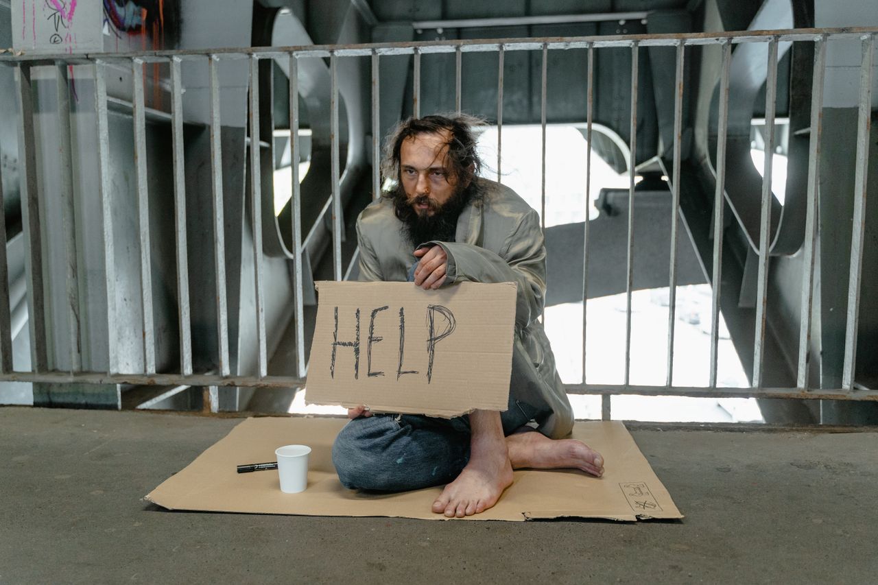 A homeless man sits under a bridge holding a 'Help' sign, highlighting poverty.